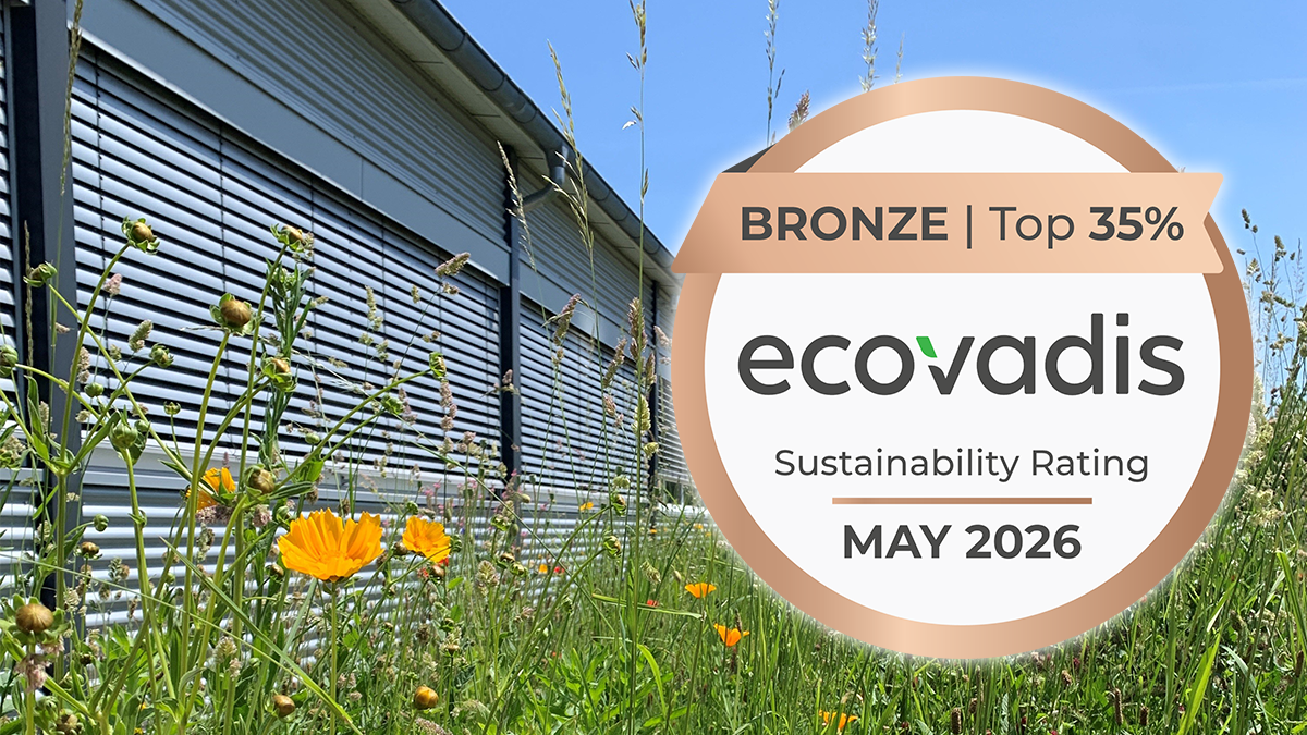 Eine bronzene EcoVadis-Nachhaltigkeitsplakette für Februar 2025 neben einem Gebäude und Wildblumen.