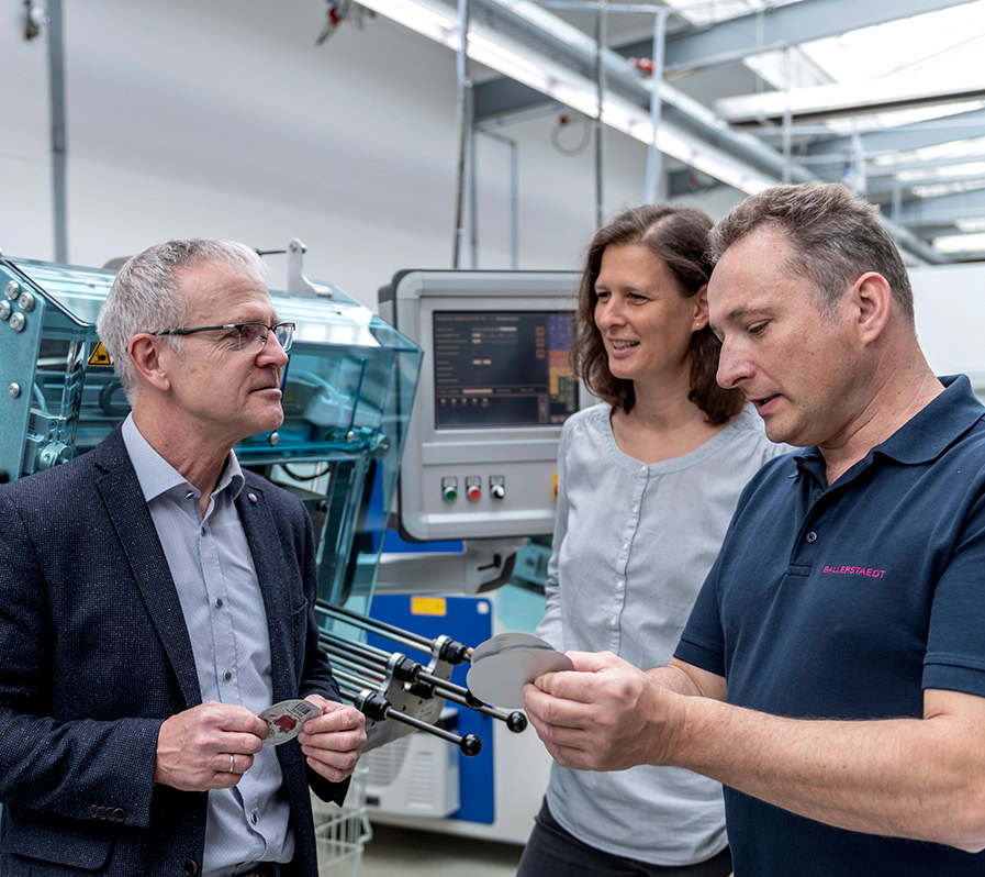Service | Beratung Drei Personen stehen vor Produktionsmaschinen. Ein Mann hält Aluminium-Siegelverschlüsse in der Hand und erklärt diese.