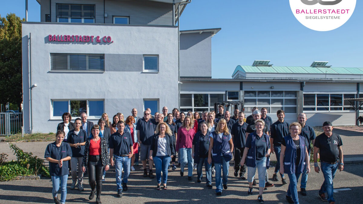 Die Belegschaft von BALLERSTAEDT, eine Gruppe von ca. 50 Menschen, steht vor dem Firmengebäude- Oben rechts im Eck ist ein Jubiläumslogo: 80 Jahre BALLERSTAEDT Siegelsysteme