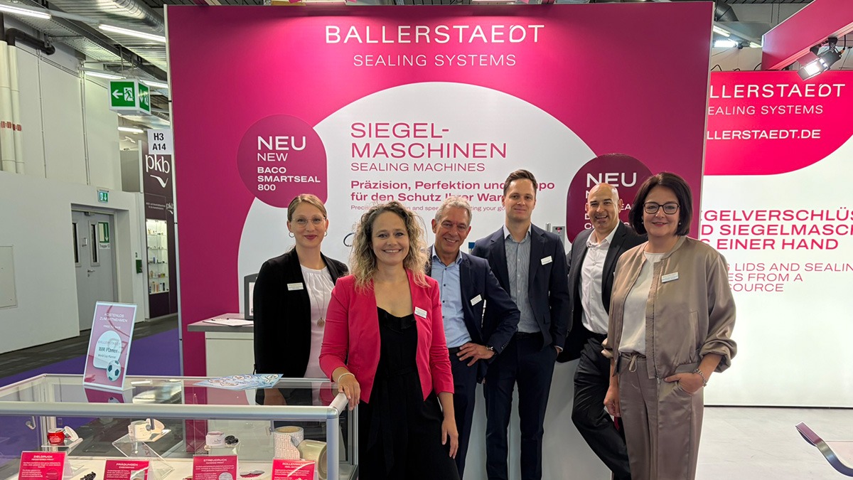 Eine Gruppe Personen steht auf einem Messestand vor einer magentafarbenen Wand. Im Vordergrund ist eine Vitrine mit versiegelten Kosmetiktiegeln zu sehen.